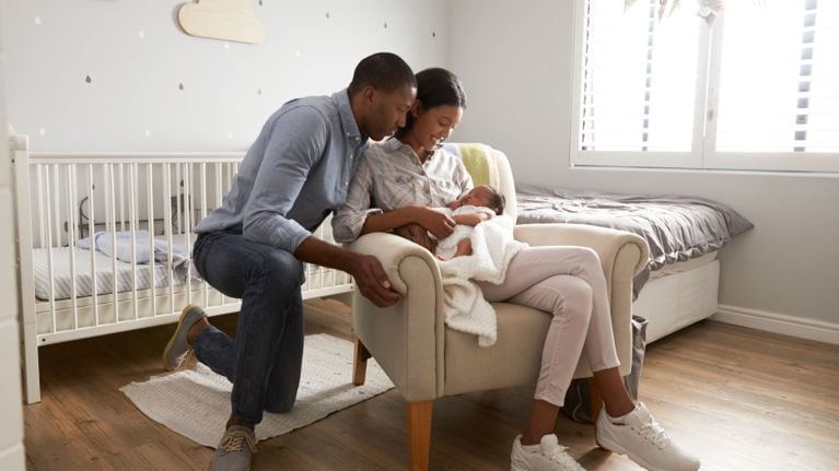 A young couple sits in a cozy nursery, admiring their sleeping baby wrapped in a white blanket.