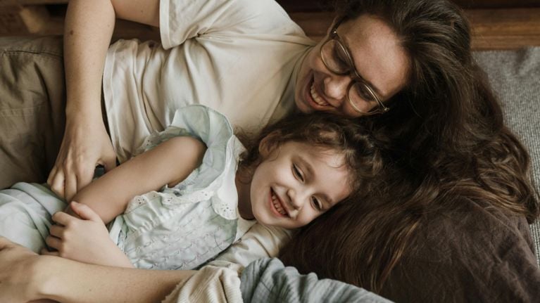 A mother and daughter are lying down together and smiling. One person is wearing glasses and the other is resting their head on the first person.