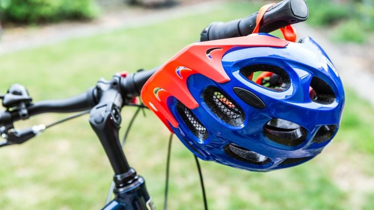 A blue bicycle helmet with a bright orange front section is hanging on bike handlebars. The helmet is positioned with the front facing towards the right.