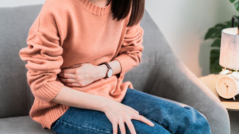 A woman in a peach sweater and jeans sits on a couch, holding her stomach with a pained expression. This image visually conveys symptoms like bloating, cramping, or digestive changes that can appear in early pregnancy.