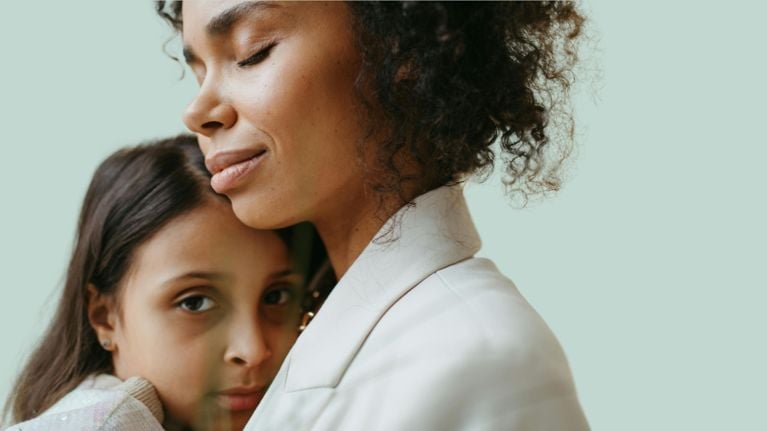 An adult is sitting on a couch, holding a young child in a hug. The adult is leaning in and kissing the child's head.