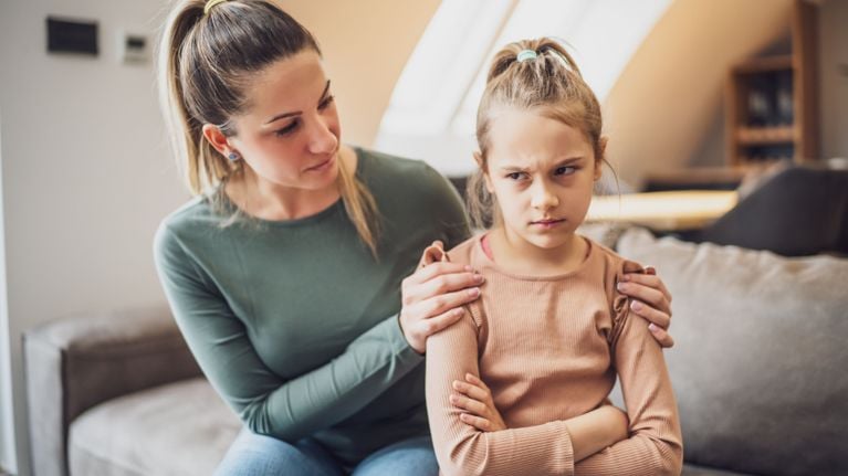 A mother with a concerned expression placing her hand gently on her daughter’s shoulder, while the child looks away with frustration and arms crossed, showing a moment of emotional tension.