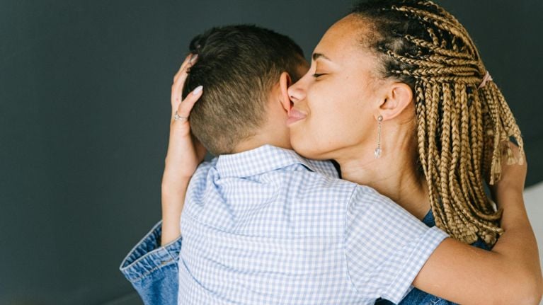 A mother and son are embracing closely, with the mother's head tilted towards her son's ear. The son has an arm around his mother's back.