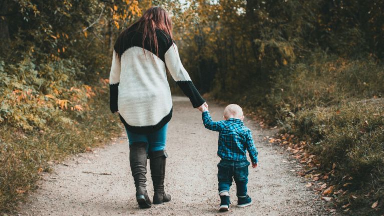 A mom and her toddler hold hands while walking a trail.