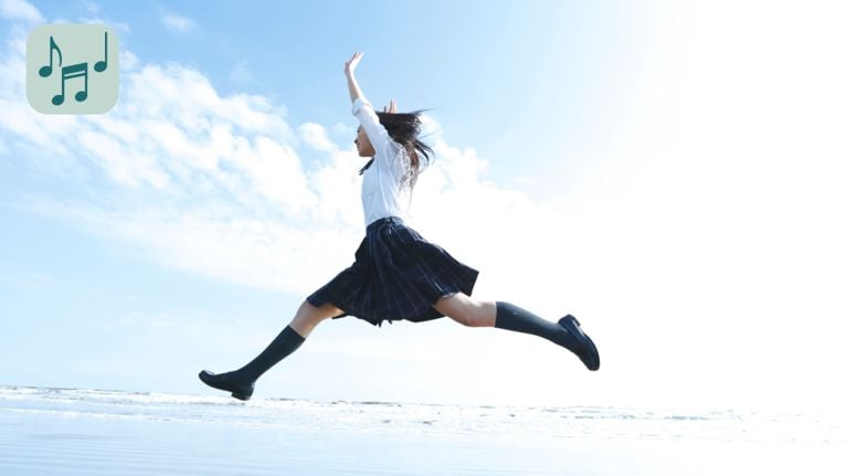 A young person is leaping into the air on a beach with their arms raised. They are a white shirt, plaid skirt, and dark socks.