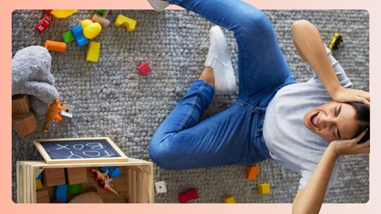 A frustrated mother sits on a carpeted floor with her hands on her head, surrounded by scattered toys and an overturned toy box. Her eyes are shut and mouth open in a scream, capturing a moment of intense overwhelm often referred to as "mom rage."