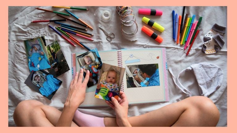 A person sitting cross-legged on a bed creates a baby scrapbook, surrounded by baby photos, colored pencils, markers, highlighters, scissors, washi tape, and baby items like socks and a hat. The open scrapbook features photos of a baby and playful illustrations.