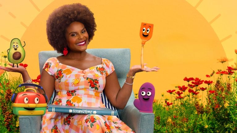 Tabitha Brown sits smiling in a sunny field of red wildflowers, balancing animated characters on each hand—including an avocado and orange spatula—while holding a “Story Time” book in her lap. The cheerful cast surrounds her in a vibrant, cartoon-style scene.