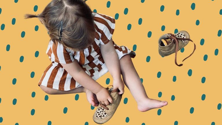 A young child wearing a striped dress sits on a dotted orange background, focused on putting on a soft, tan shoe with leopard-print detailing.