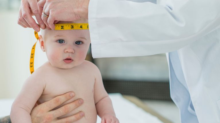 A healthcare professional uses a yellow measuring tape to check an infant's head circumference during a check-up.