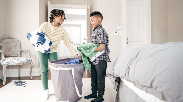 Two children pull towels out of a grey laundry hamper.