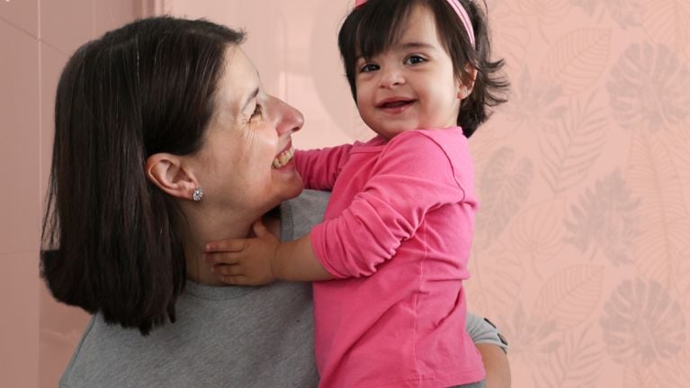 Mother in her 40s holding her happy baby girl, both smiling warmly against a soft pink background, highlighting connection and contentment.