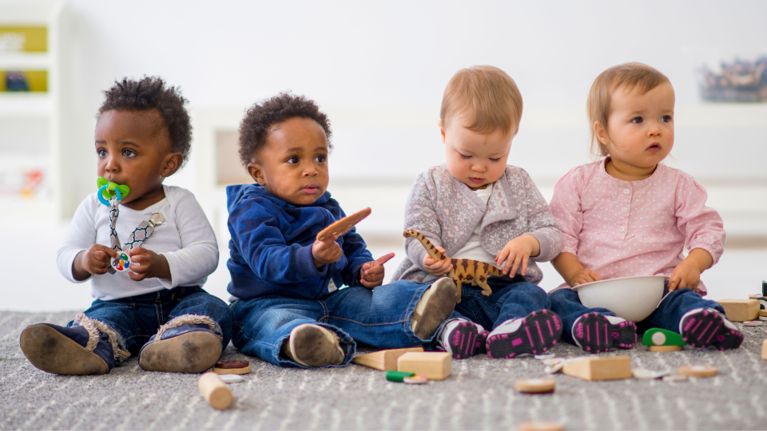 Four toddlers sit in a row on the floor playing with toys against a bright, clean indoor backdrop.