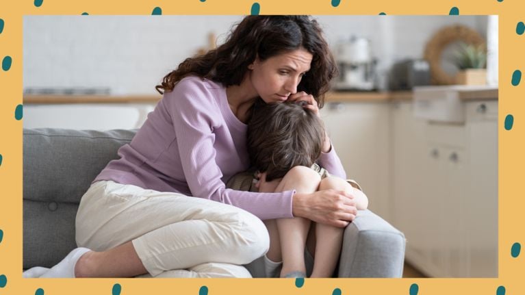 A woman sits on a couch hugging a young child who is curled up and appears distressed. The woman looks concerned and is gently holding the child close. They are in a cozy kitchen or living space.