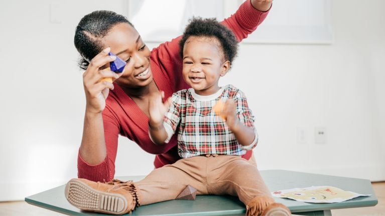 A smiling toddler sits on a table next to a woman, both playing with colorful blocks and toys in a well-lit
