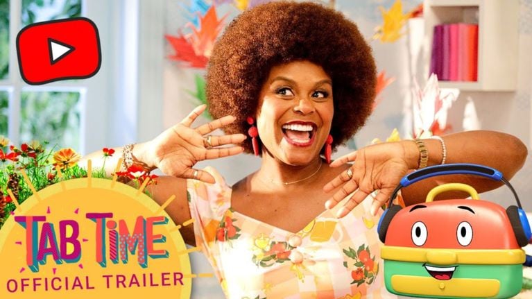 Tabitha Brown beams joyfully with her hands raised mid-dance. She wears a fruit-printed dress beside a smiling animated lunchbox with headphones. A YouTube play button hovers above, and “Tab Time Official Trailer” is written in a sunburst design.