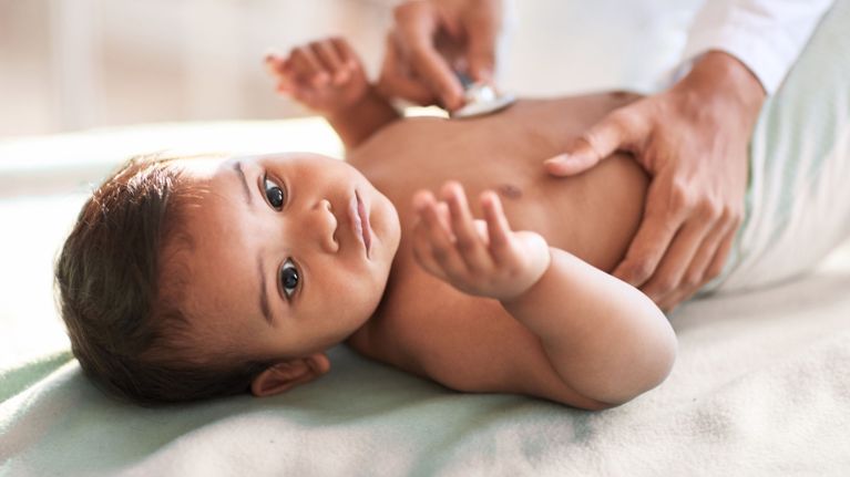 A baby looks up while lying on an exam table as a doctor listens to their chest with a stethoscope during a wellness check.