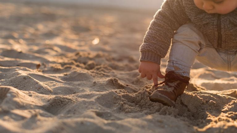 A toddler crouches on a sandy beach at sunset, pointing at their brown boot while dressed in a cozy sweater and jeans.