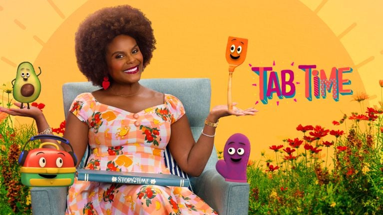 Tabitha Brown sits in a garden chair with a “Story Time” book across her lap, smiling as animated friends perch on each hand. A sunny orange backdrop highlights the “Tab Time” logo on the right, surrounded by blooming red flowers.