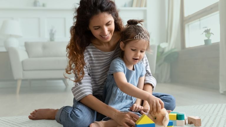 A woman sits on the floor with a young girl who wears a crown. They are playing with blocks.