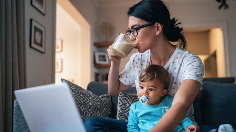 A parent multitasking at home—sitting on the couch with a toddler while drinking coffee and looking at a laptop—symbolizing the challenge of staying informed about child safety recalls.