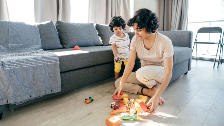 A mother kneels on the floor beside her toddler, examining a pile of colorful toys in a living room. The child watches closely, holding a small car, while natural light fills the space through large windows.
