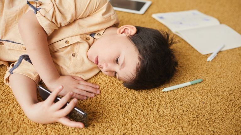 A boy lying on his side on a yellow carpet and looking at his phone