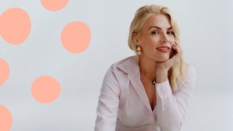 A woman, Busy Philipps, rests her chin in her cheek in a colourful pink outfit surrounded by painting equipment