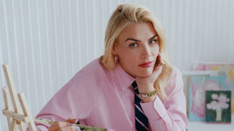 A woman, Busy Philipps, rests her chin in her cheek in a colourful pink outfit surrounded by painting equipment