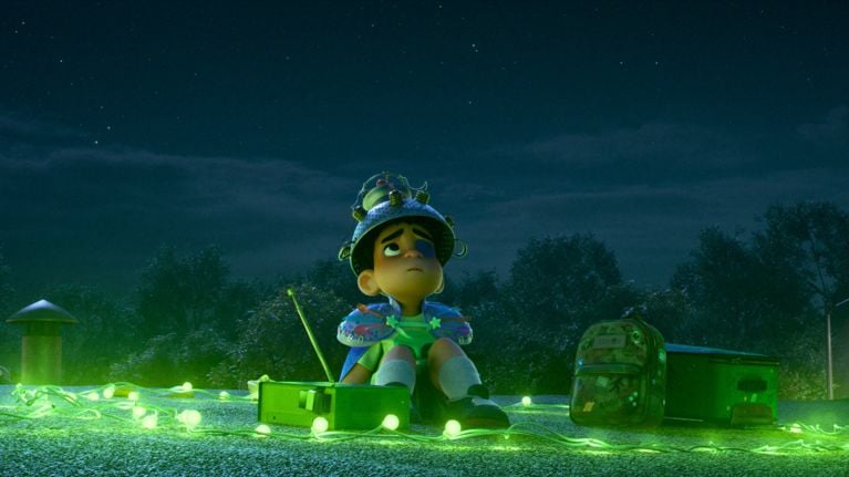 A still from the animated movie Elio showing the main character sitting alone surrounded by green lights