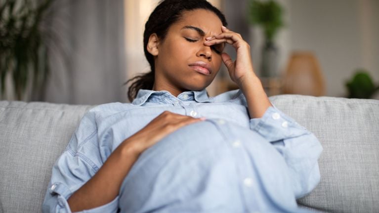 A pregnant person reclines on a couch, eyes closed and hand to forehead in discomfort—illustrating sinus pressure or nasal symptoms often experienced during pregnancy.