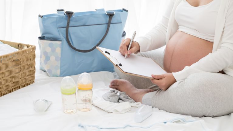 A pregnant woman with a diaper bag and two baby bottles writing something on a clipboard