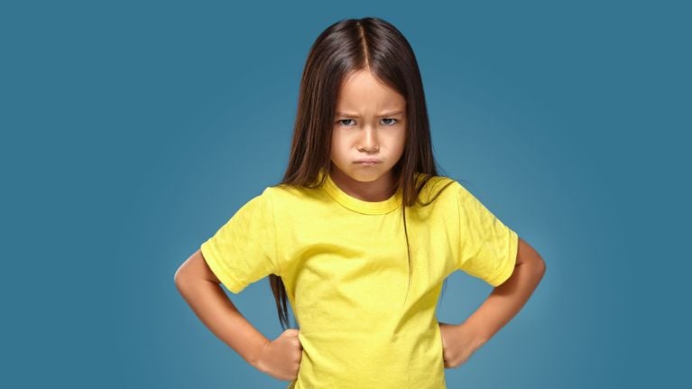 Young girl with long brown hair wearing a bright yellow T-shirt, standing with hands on hips and a serious, angry expression against a solid blue background.