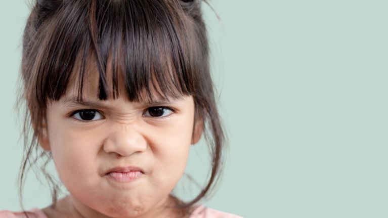 Young girl with long brown hair wearing a bright yellow T-shirt, standing with hands on hips and a serious, angry expression against a solid blue background.