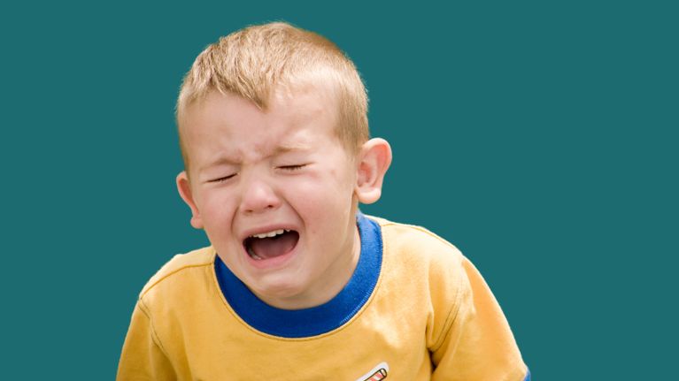 Young boy with short blond hair wearing a yellow shirt with a blue collar, crying with eyes closed and mouth open against a solid teal background.