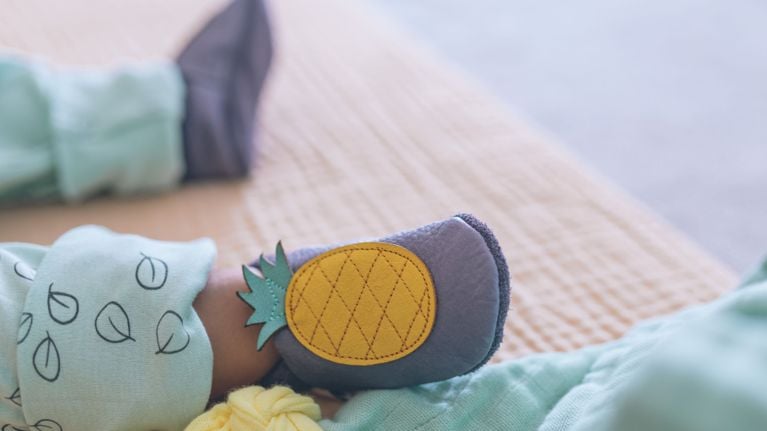 A close-up of a baby wearing soft grey baby shoes with a yellow pineapple design on top, lying on a bed with a light quilted blanket. The baby’s outfit includes mint green pants and a onesie with black leaf prints.