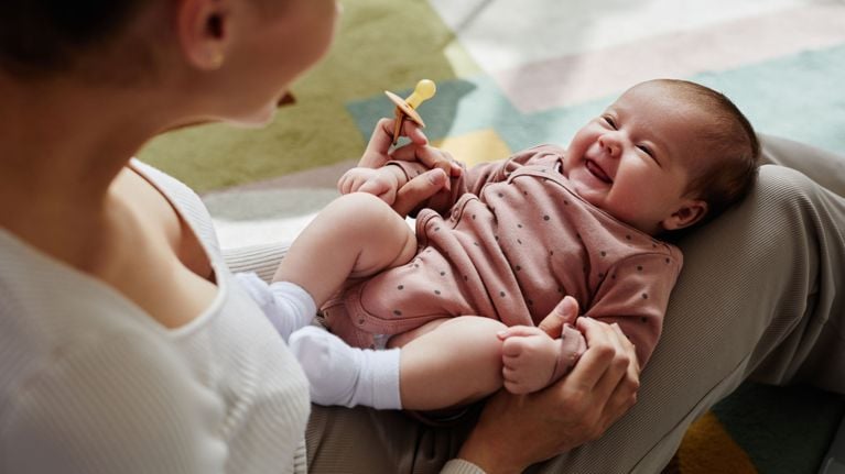 A smiling baby in a pink polka-dot onesie lies on a caregiver’s lap, holding a pacifier and looking up joyfully. The caregiver, partially visible, engages warmly with the infant in a cozy, carpeted space. The image captures bonding and early social connection.
