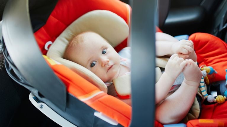 A baby with light hair and blue eyes sitting in a rear-facing infant car seat with a red and beige interior, looking alert and curious.