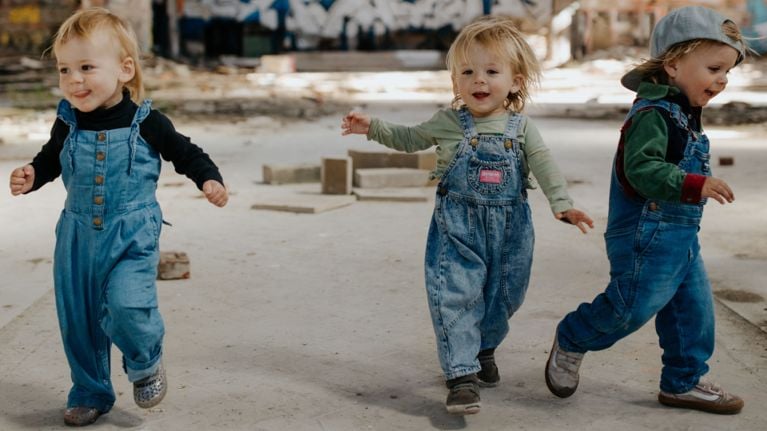 Three toddlers in denim overalls joyfully run and play together in a sunlit, industrial-style space. The children, all with light hair, appear carefree and full of energy, surrounded by scattered blocks and a graffiti-covered wall in the background.