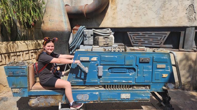 Teen girl wearing Star Wars Mickey ears sits on a blue, sci-fi speeder-style vehicle in Galaxy’s Edge at Disneyland, smiling in the sun with themed props and tubing behind her.