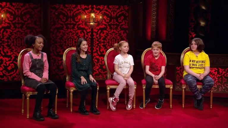 Five children sit in a row of ornate red and gold chairs against a red damask-patterned backdrop on the set of Junior Taskmaster. Each child wears bright, expressive clothing, smiling and looking engaged as they await their task or feedback.