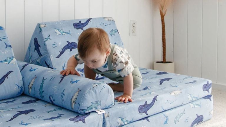 A baby crawls on a Nugget couch covered in blue upholstery with a whale and shark pattern, in a white-paneled room with soft carpet and a tall floor plant.