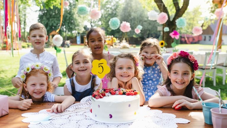 A group of smiling children gathered around a birthday cake at an outdoor party. The cake features a yellow “5” topper, and the background is decorated with hanging ribbons and pastel tissue paper pom-poms.
