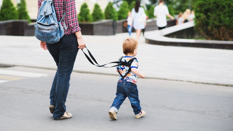 A young child in a checkered shirt, beanie, and jeans looks toward the camera while walking on a busy pedestrian path. The child is secured with a black chest-style safety harness held by an adult just behind them.