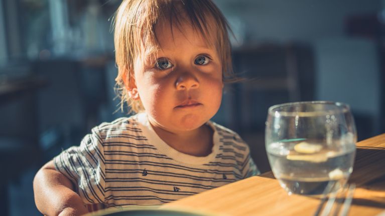 A toddler with light brown hair and a serious expression sits at a wooden table in a sunlit room. A clear glass of water with lemon slices is placed nearby. The image suggests quiet, screen-free mealtime or reflective moments during eating.