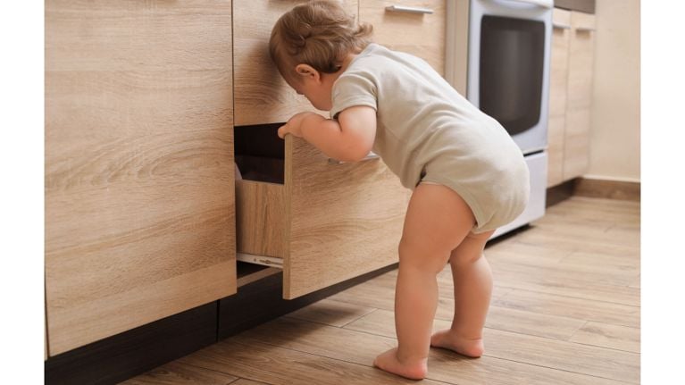 A curious toddler in a beige onesie stands in a modern kitchen, peering into an open lower drawer. The image highlights the importance of childproofing drawers and cabinets to keep toddlers safe at home.