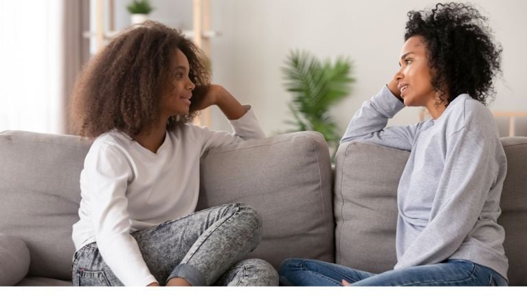 A teenage girl and her mother sit on a grey couch, smiling and making eye contact as they talk openly at home.