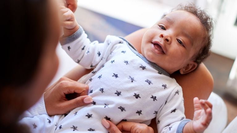 A baby wearing a white onesie with black stars smiles and gazes upward while being held by a caregiver. The baby’s hands are mid-motion, suggesting playful interaction and emotional engagement during a quiet bonding moment.