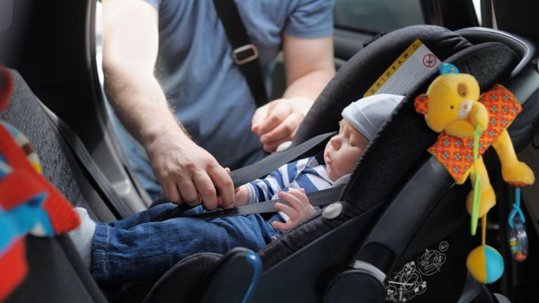An adult securing a sleeping baby into a black rear-facing car seat using the harness straps, with a yellow stuffed bear toy hanging from the handle.