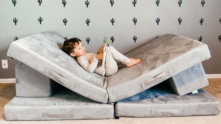 A young child lounges on a grey Figgy couch arranged into a reclined reading nook, holding a book in a room with cactus-print wallpaper.
